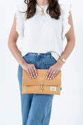 A woman wearing a white ruffle-sleeve top and blue jeans holds a tan leather clutch bag with a gold clasp against a plain white background. Handmade by Meanwhile Back on the Farm in Lynchburg,VA