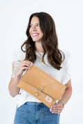 A woman with long brown hair smiling and looking to the side while holding a tan leather clutch bag with a gold clasp. Handmade by Meanwhile Back on the Farm in Lynchburg,VA