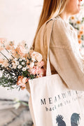 Girl wearing a beige Meanwhile back on the farm tote bag with the black rooster logo with flowers inside