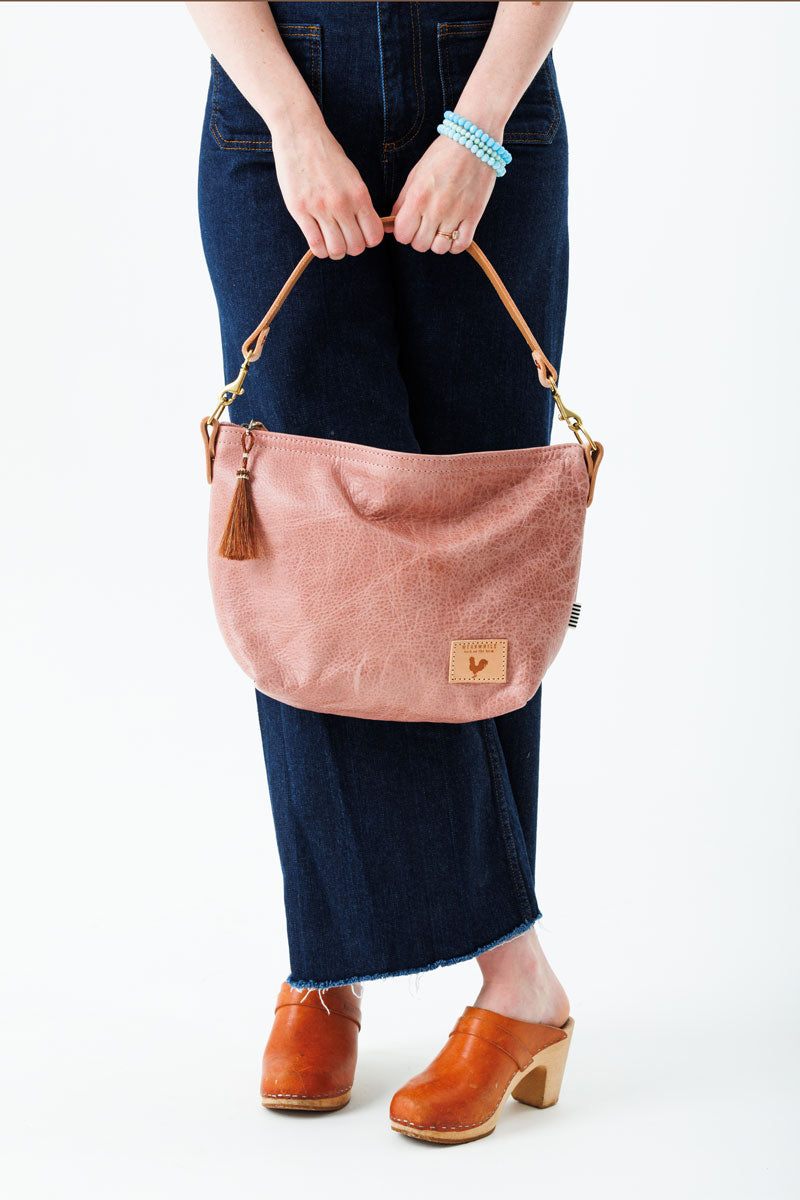 A woman wearing over the shoulder the rose pink bag with the meanwhile logo and a brown tassel zipper.