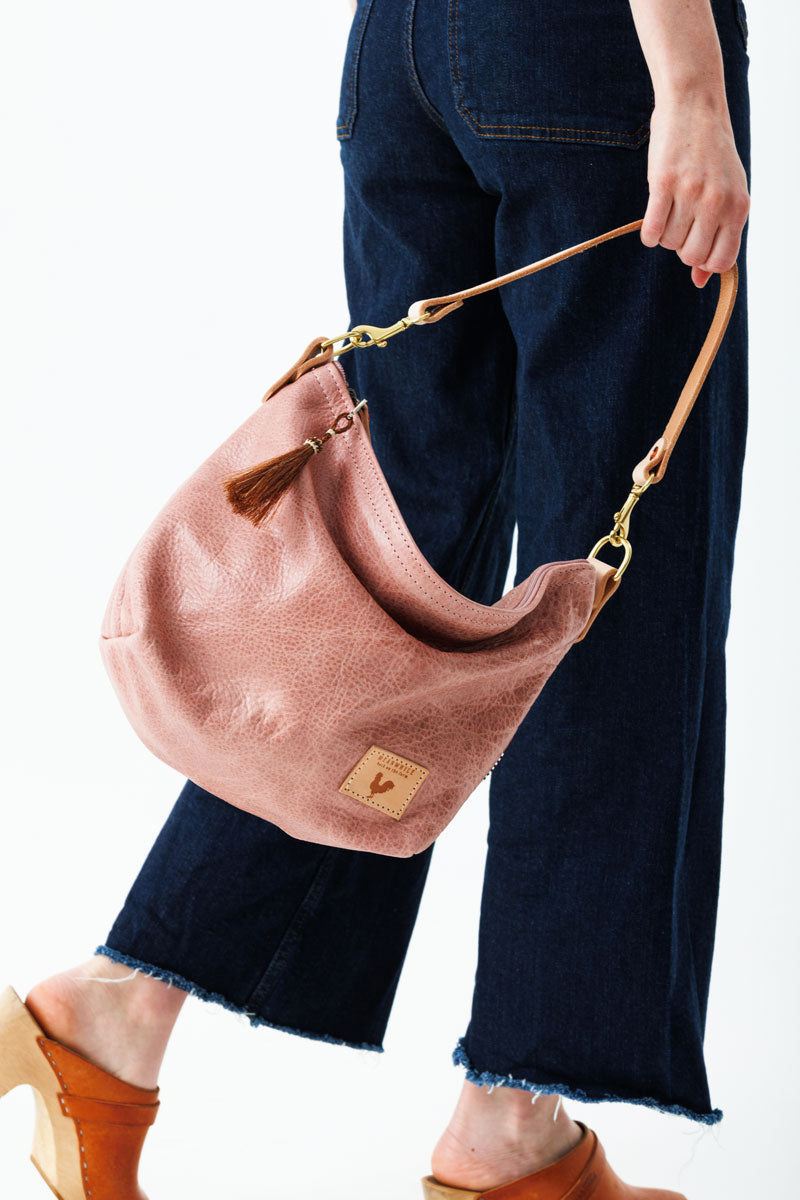 A woman wearing over the shoulder the rose pink bag with the meanwhile logo and a brown tassel zipper.