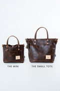 Two dark brown leather tote bags shown side by side on a light gray background, labeled 'THE MINI' and 'THE SMALL TOTE'. The larger bag on the right has double top handles, a front pocket, and a tassel detail, while the smaller bag on the left is a compact matching tote. Handmade by Meanwhile Back on the Farm in Lynchburg,VA