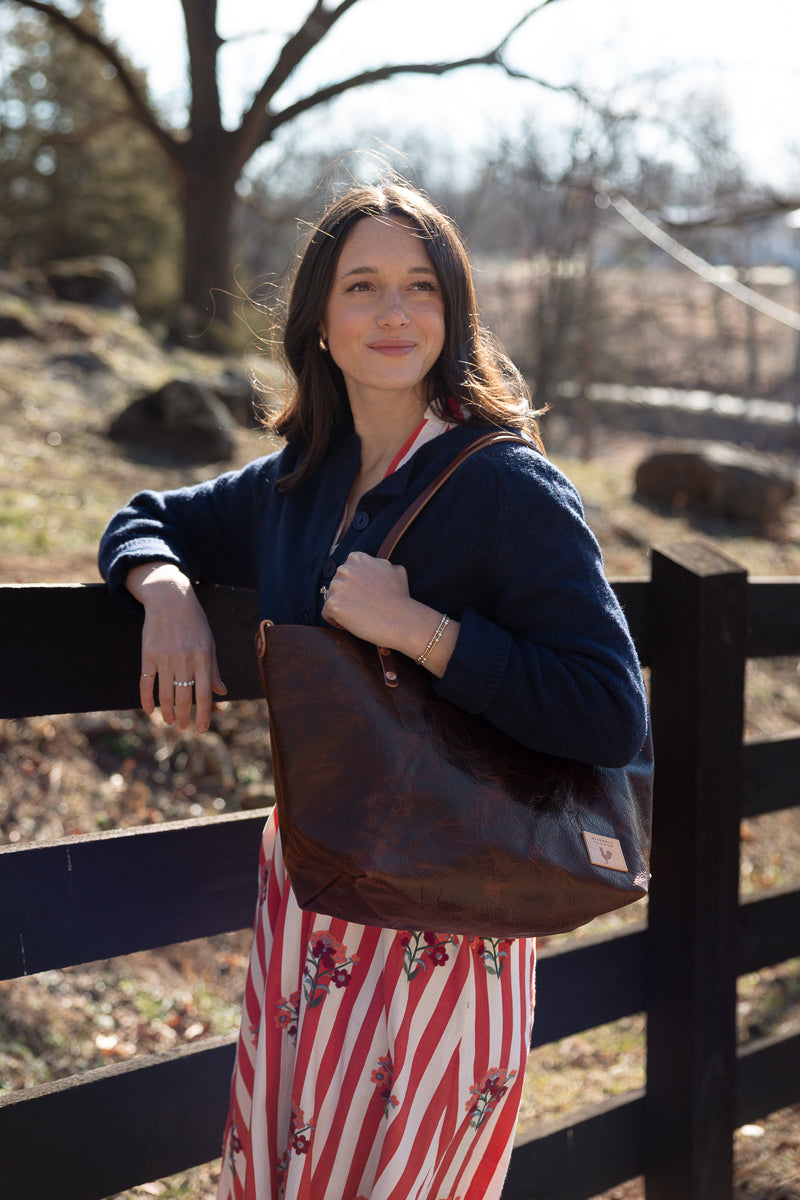 A woman wearing a navy cardigan and a red-and-white striped skirt with floral details, carrying a large brown leather shoulder bag, leaning on a dark wooden fence in an outdoor setting. Handcrafted by Meanwhile Back on the Farm in Lynchburg, Va.