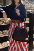 A woman outdoors wearing a navy cardigan, a red and white striped skirt with a floral print, and a brown belt, carrying a dark brown tote bag. Handcrafted by Meanwhile Back on the Farm in Lynchburg, Va.