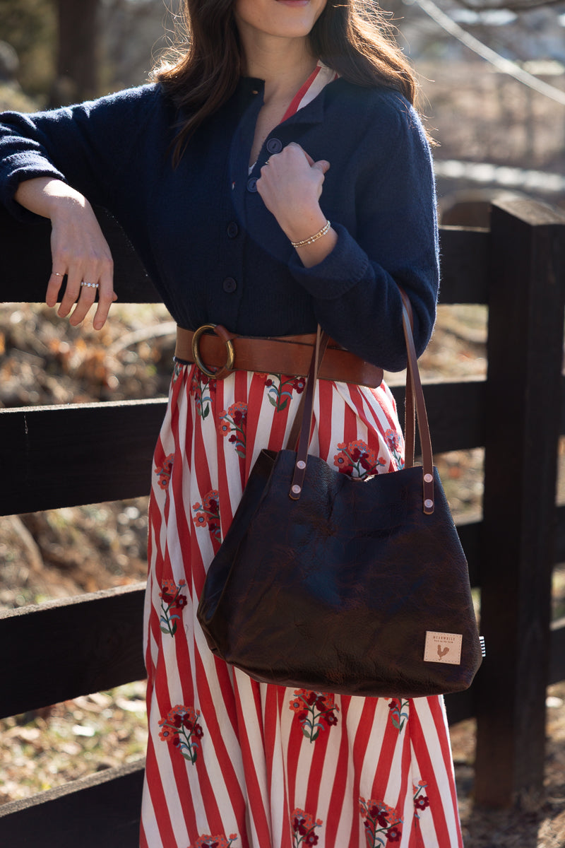 A woman outdoors wearing a navy cardigan, a red and white striped skirt with a floral print, and a brown belt, carrying a dark brown tote bag. Handcrafted by Meanwhile Back on the Farm in Lynchburg, Va.