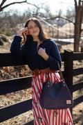 A woman outdoors leaning against a dark wooden fence, wearing a navy long-sleeve button-up shirt, a red and white vertically striped skirt with a floral print, a brown belt, and carrying a dark navy tote bag. Handcrafted by Meanwhile Back on the Farm in Lynchburg, Va.