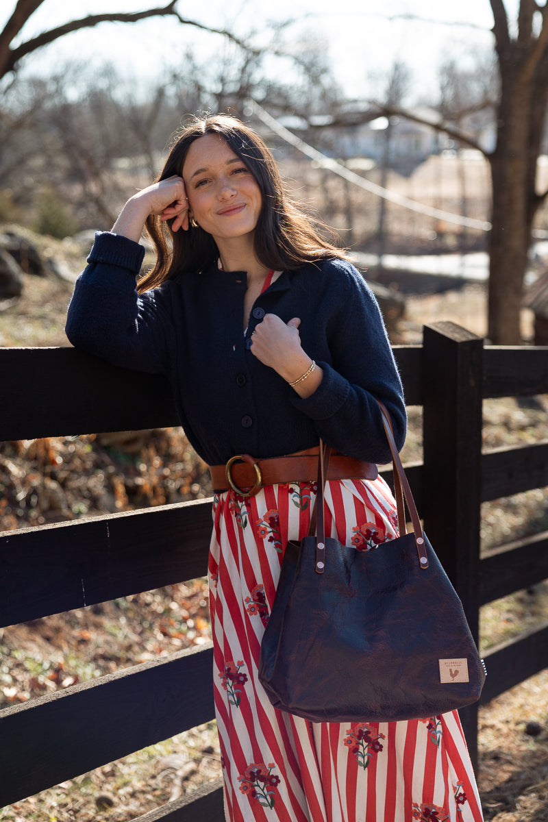 A woman outdoors leaning against a dark wooden fence, wearing a navy long-sleeve button-up shirt, a red and white vertically striped skirt with a floral print, a brown belt, and carrying a dark navy tote bag. Handcrafted by Meanwhile Back on the Farm in Lynchburg, Va.