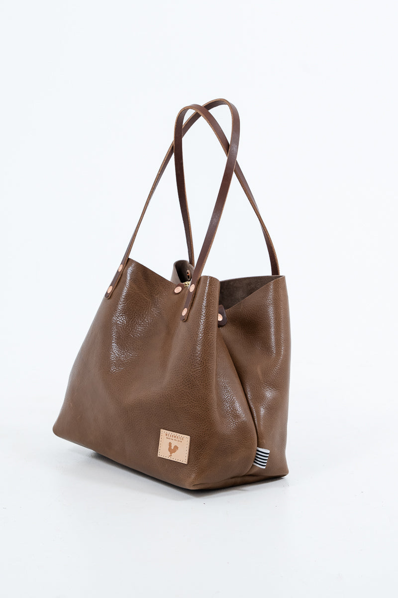 A chestnut brown leather handbag with a visible brand logo on a white background. Handmade by Meanwhile Back on the Farm in Lynchburg,VA background