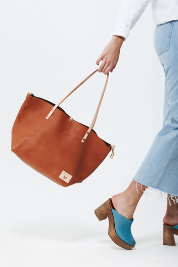 Model casually swinging medium sized saddle brown leather tote with cream colored shoulder straps.