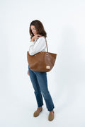 A woman wearing a white ribbed long-sleeve top, blue jeans, and tan loafers carries a large chestnut brown leather tote bag against a plain white studio background.  Handmade by Meanwhile Back on the Farm in Lynchburg, Va.