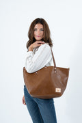 A woman wearing a white long-sleeve knit top and blue jeans carries a large chestnut brown leather tote bag over her shoulder against a plain white studio background.  Handmade by Meanwhile Back on the Farm in Lynchburg, Va.