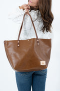 A woman wearing a white knit top and blue jeans holds up a large chestnut brown leather tote bag with double shoulder straps and a small beige brand patch on the bottom right corner.  Handmade by Meanwhile Back on the Farm in Lynchburg, Va.