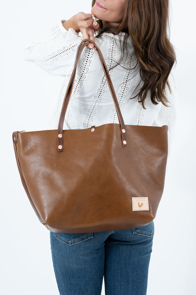 A woman wearing a white knit top and blue jeans holds up a large chestnut brown leather tote bag with double shoulder straps and a small beige brand patch on the bottom right corner.  Handmade by Meanwhile Back on the Farm in Lynchburg, Va.
