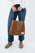 A person wearing blue jeans and brown shoes holds a large chestnut brown leather tote bag with pearl-like hardware and a small tan brand patch. The photo is cropped from the chest down on a clean white studio background.  Handmade by Meanwhile Back on the Farm in Lynchburg, Va.