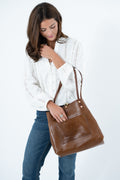 A woman wearing a white eyelet blouse and blue jeans holds a large walnut brown leather tote bag against a plain white studio background. Handmade by Meanwhile Back on the Farm in Lynchburg, Va.