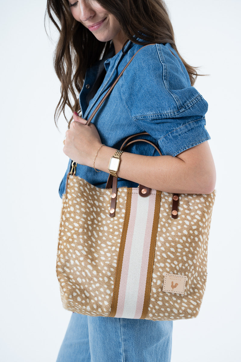 A woman in a blue denim shirt and jeans carries a large tote bag with a tan and white spotted pattern, brown leather handles, and vertical brown and white stripes on the front. Proudly handcrafted by Meanwhile Back on the Farm in Lynchburg, Va.