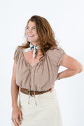A woman with wavy light brown hair smiling and looking to the side, wearing a brown and beige gingham short-sleeve blouse with a light floral neck scarf, a brown belt, and cream-colored pants, set against a plain white background. Proudly handcrafted by Meanwhile Back on the Farm in Lynchburg, Va.