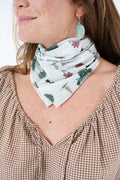 A close-up of a woman wearing a white neck scarf with a colorful topiary and floral print, styled with a brown and white gingham top. Proudly handcrafted by Meanwhile Back on the Farm in Lynchburg, Va.