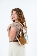 A woman with shoulder-length brown hair is shown from the back and side, wearing a brown leather backpack with a floral scarf tucked into the side pocket. She is wearing a brown and white gingham top and cream-colored pants, set against a plain white studio background. Proudly handcrafted by Meanwhile Back on the Farm in Lynchburg, Va.