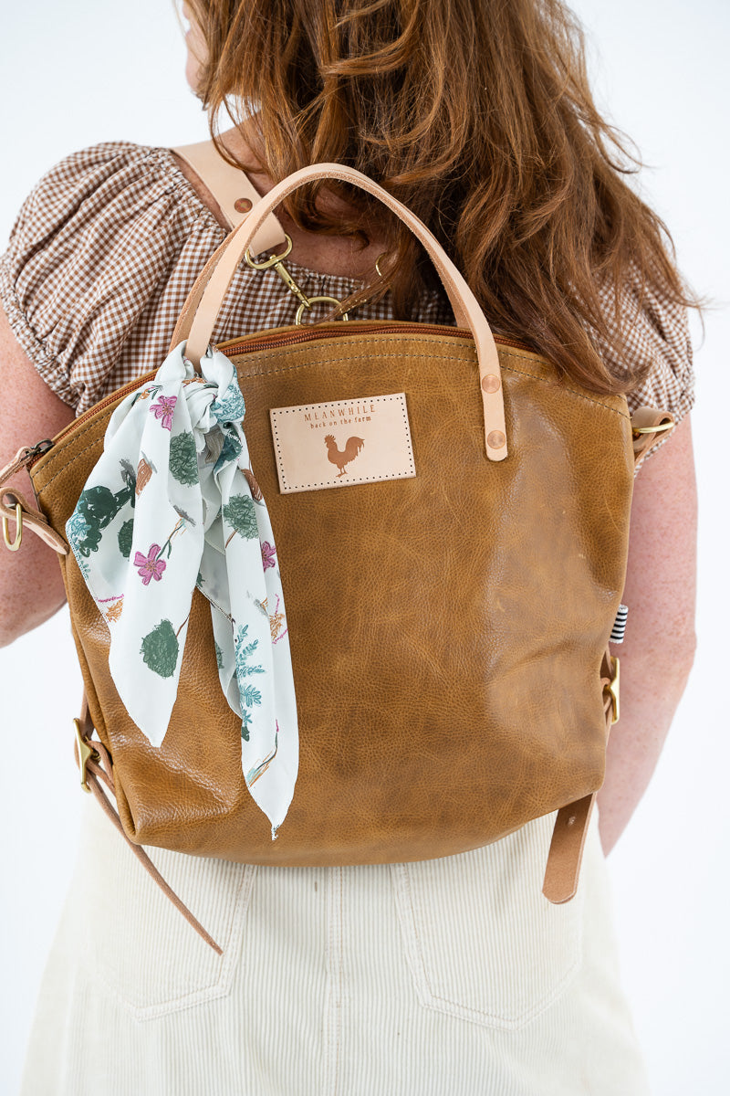 A woman viewed from behind wearing a tan leather tote backpack with a floral-printed scarf hanging from the handle. The backpack has a rectangular leather patch on the front with a rooster logo and the text 'Meanwhile Back on the Farm'. Proudly handcrafted by Meanwhile Back on the Farm in Lynchburg, Va.