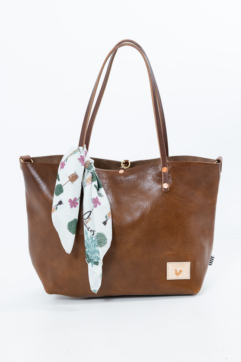 A brown leather tote bag with dual shoulder straps, a gold-tone clasp closure, and a small beige brand patch on the lower right. A white silk scarf with a green and pink floral print is tied to the left side of the bag. Proudly handcrafted by Meanwhile Back on the Farm in Lynchburg, Va.