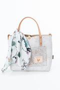 A white textured tote bag with light brown leather handles, featuring a matching white scarf with a green and pink botanical print tied to the handle. The bag has a front pocket with a tan brand patch and is set against a plain white background. Proudly handcrafted by Meanwhile Back on the Farm in Lynchburg, Va.