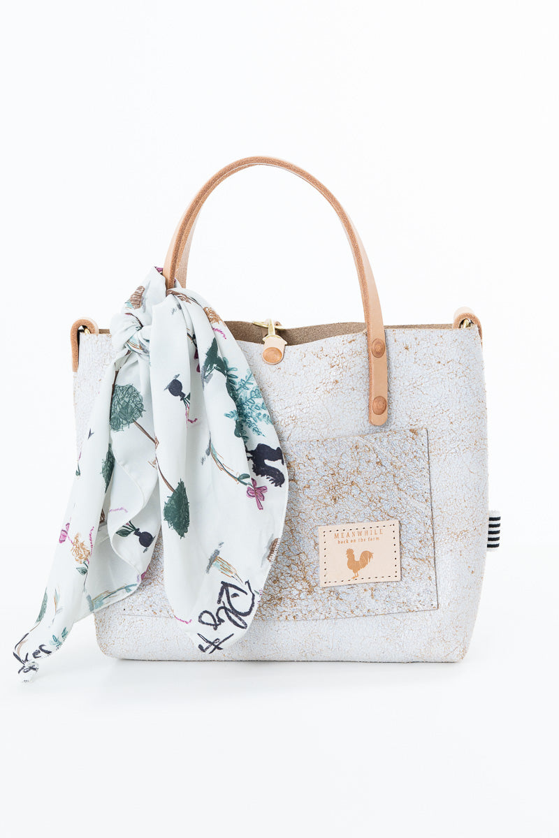 A white textured tote bag with light brown leather handles, featuring a matching white scarf with a green and pink botanical print tied to the handle. The bag has a front pocket with a tan brand patch and is set against a plain white background. Proudly handcrafted by Meanwhile Back on the Farm in Lynchburg, Va.