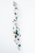A white knotted scarf featuring a whimsical pattern of potted cacti, succulents, and small flowers in green, brown, and pink, set against a plain white background. Proudly handcrafted by Meanwhile Back on the Farm in Lynchburg, Va.