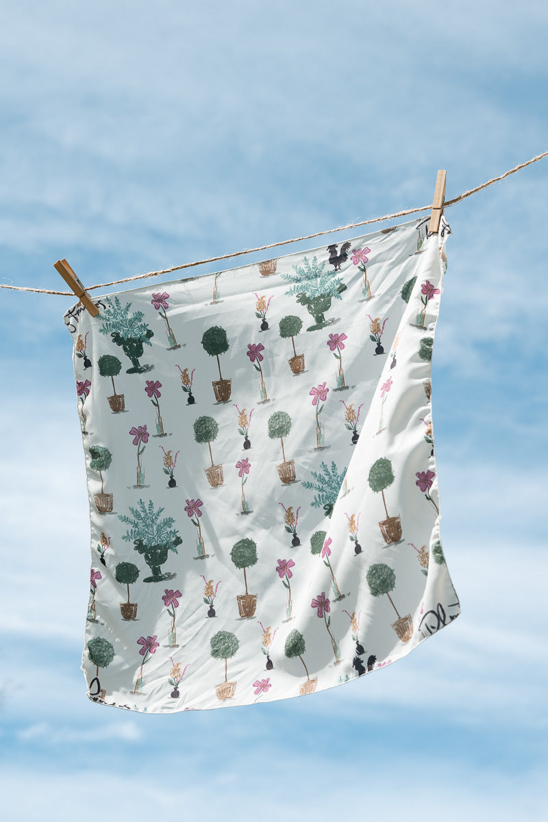 A white fabric cloth with a colorful pattern of potted plants hanging from a clothesline with wooden clips against a blue sky background. Proudly handcrafted by Meanwhile Back on the Farm in Lynchburg, Va.