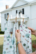 A person's hands hang a white fabric garment with a botanical tree print on a clothesline in front of a white house. Proudly handcrafted by Meanwhile Back on the Farm in Lynchburg, Va.