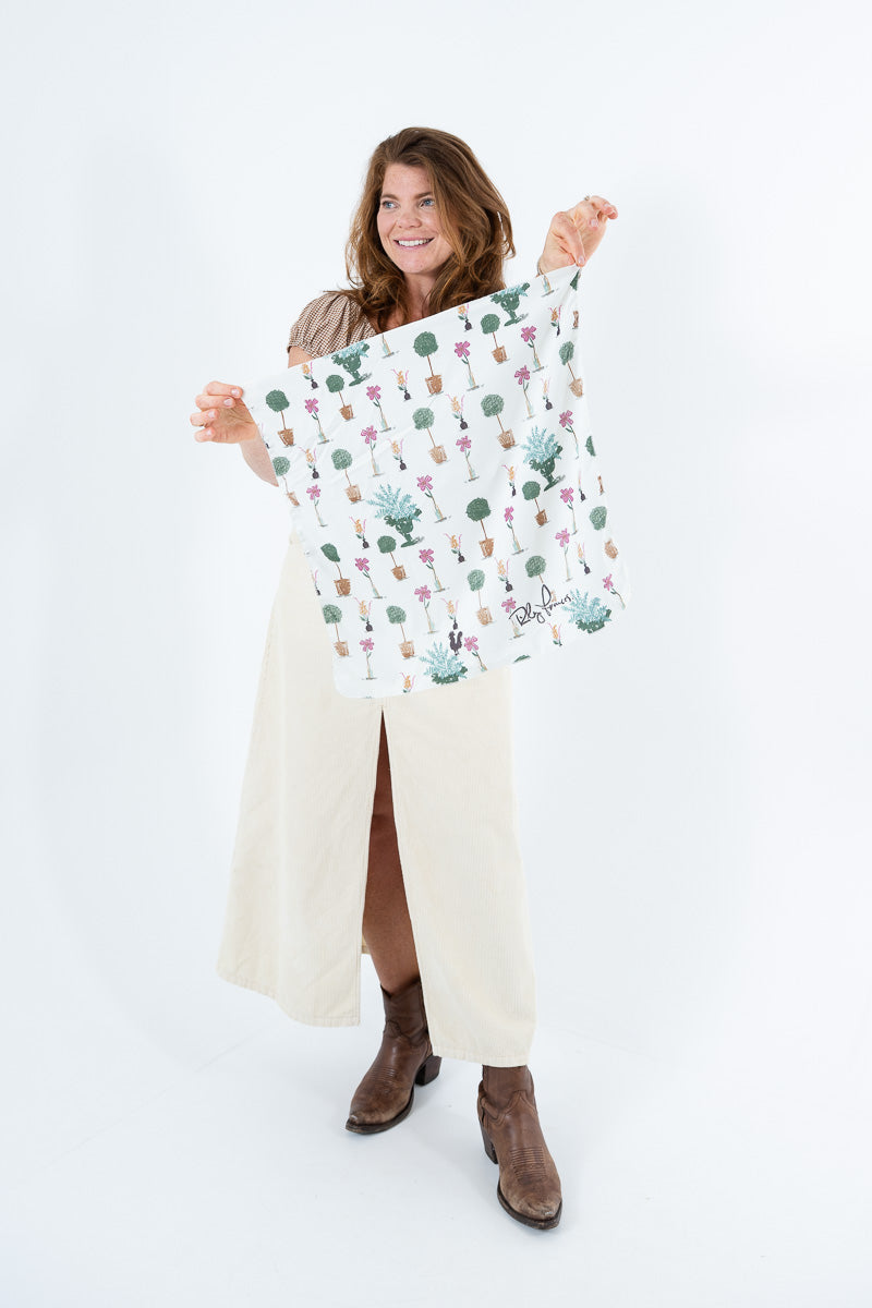 A woman stands against a white studio background holding up a square scarf or bandana with a colorful cactus and succulent pattern. She is wearing a beige dress and brown ankle boots. Proudly handcrafted by Meanwhile Back on the Farm in Lynchburg, Va.