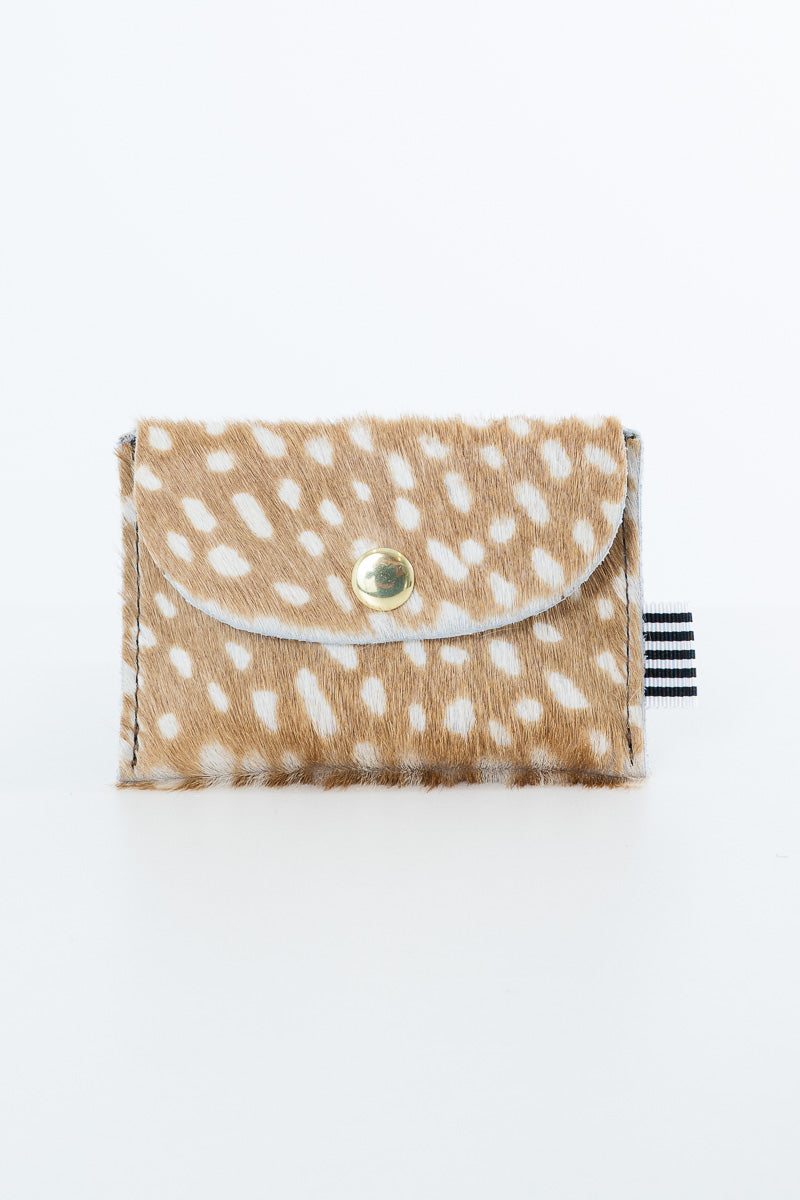 A small bifold wallet made of tan and white spotted faux fur or hair-on-hide material, with a flap closure and a gold snap button. A small black-and-white striped fabric tag is visible on the right side. Proudly handcrafted by Meanwhile Back on the Farm in Lynchburg, Va.