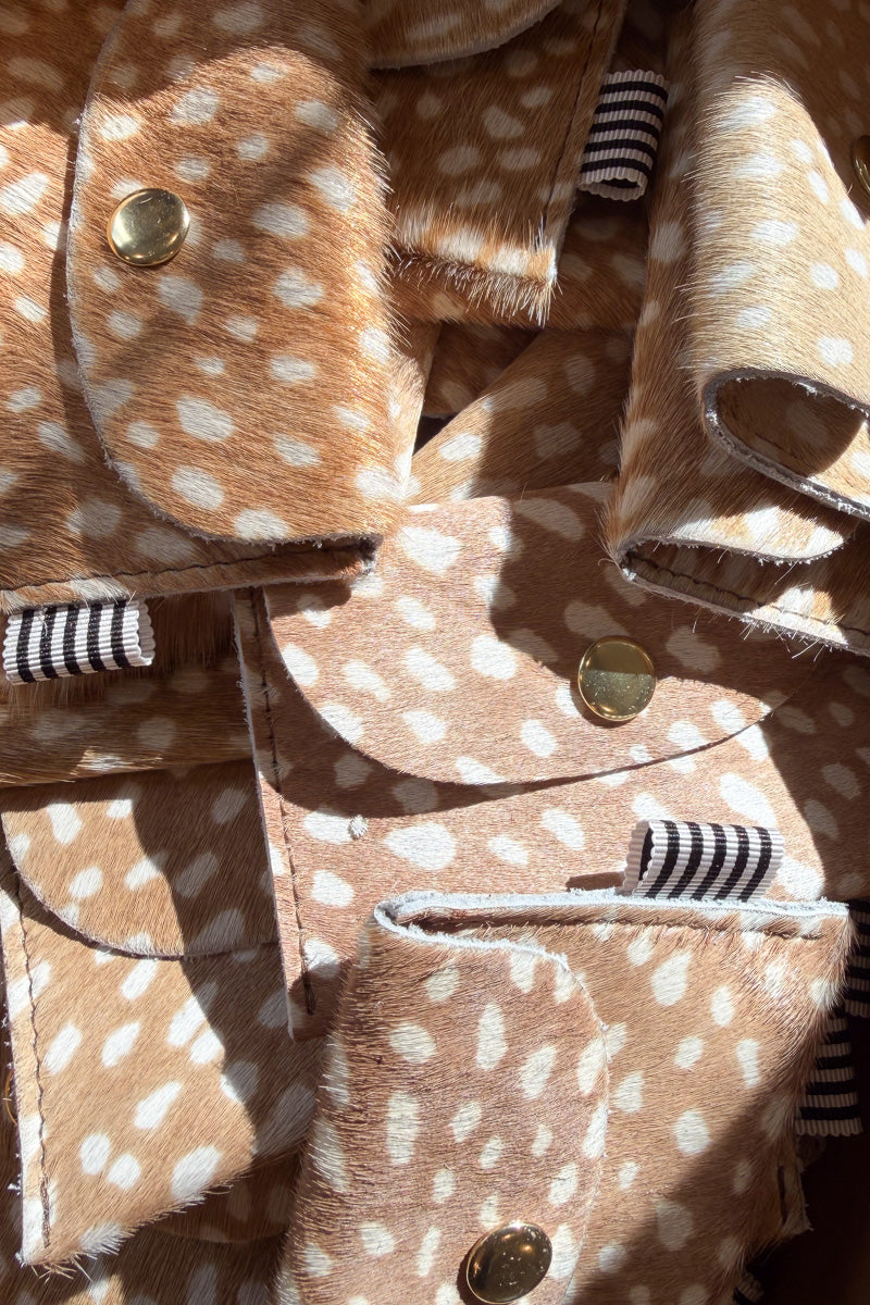 A small bifold wallet made of tan and white spotted faux fur or hair-on-hide material, with a flap closure and a gold snap button. A small black-and-white striped fabric tag is visible on the right side. Proudly handcrafted by Meanwhile Back on the Farm in Lynchburg, Va.