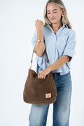 A woman wearing a light blue button-up shirt and blue jeans carries a brown suede bucket bag with a brown leather strap and a small tan logo patch on the front, set against a plain white studio background. Handcrafted by Meanwhile Back on the Farm in Lynchburg, VA.