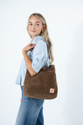 A woman wearing a light blue button-up shirt and blue jeans carries a large brown suede shoulder bag with a small tan leather logo patch, set against a plain white studio background. Handcrafted by Meanwhile Back on the Farm in Lynchburg, VA.