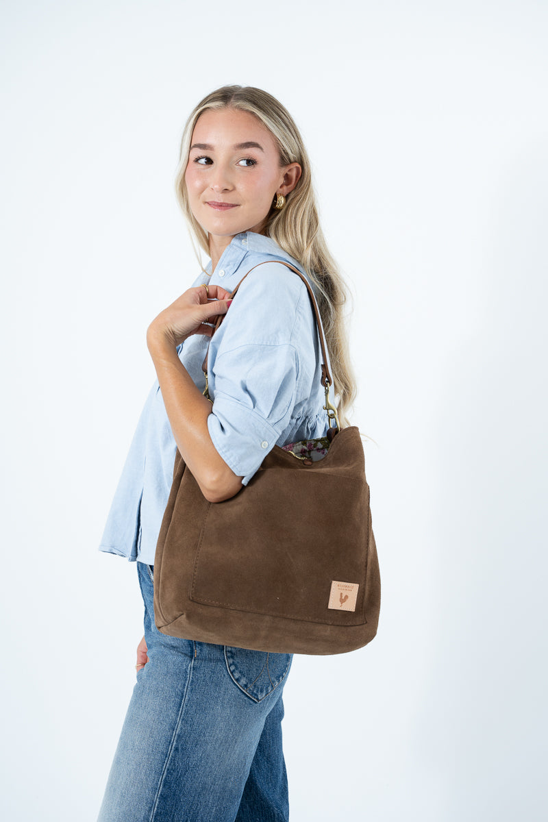 A woman wearing a light blue button-up shirt and blue jeans carries a large brown suede shoulder bag with a small tan leather logo patch, set against a plain white studio background. Handcrafted by Meanwhile Back on the Farm in Lynchburg, VA.