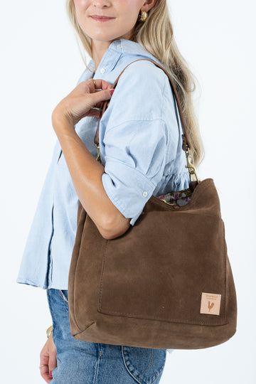 A woman wearing a light blue button-up shirt and jeans carries a large brown suede shoulder bag with a small tan brand patch on the bottom right corner, set against a plain white background. Handcrafted by Meanwhile Back on the Farm in Lynchburg, VA.
