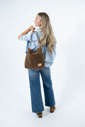 A woman with long blonde hair is shown from the back wearing a light blue denim shirt and wide-leg blue jeans, carrying a brown suede shoulder bag. She is standing against a clean white studio background. Handcrafted by Meanwhile Back on the Farm in Lynchburg, VA.