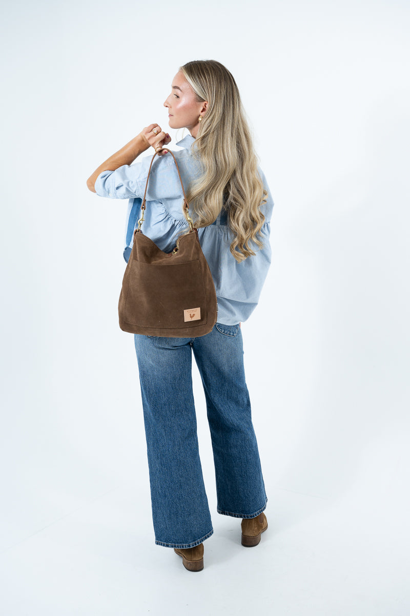 A woman with long blonde hair is shown from the back wearing a light blue denim shirt and wide-leg blue jeans, carrying a brown suede shoulder bag. She is standing against a clean white studio background. Handcrafted by Meanwhile Back on the Farm in Lynchburg, VA.