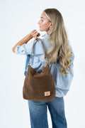 A woman wearing a light blue shirt and jeans carries a brown suede shoulder bag with a tan leather patch on a plain white background. Handcrafted by Meanwhile Back on the Farm in Lynchburg, VA.