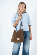 A woman wearing a light blue shirt and blue jeans carries a brown suede shoulder bag with a brown leather strap and a small tan logo patch on the front, set against a plain white studio  background. Handcrafted by Meanwhile Back on the Farm in Lynchburg, VA.
