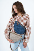 A woman wearing a brown and white gingham shirt and light blue jeans models two textured crescent-shaped crossbody bags, one in blue and one in taupe, against a plain white background. Handcrafted by Meanwhile Back on the Farm in Lynchburg, Va.