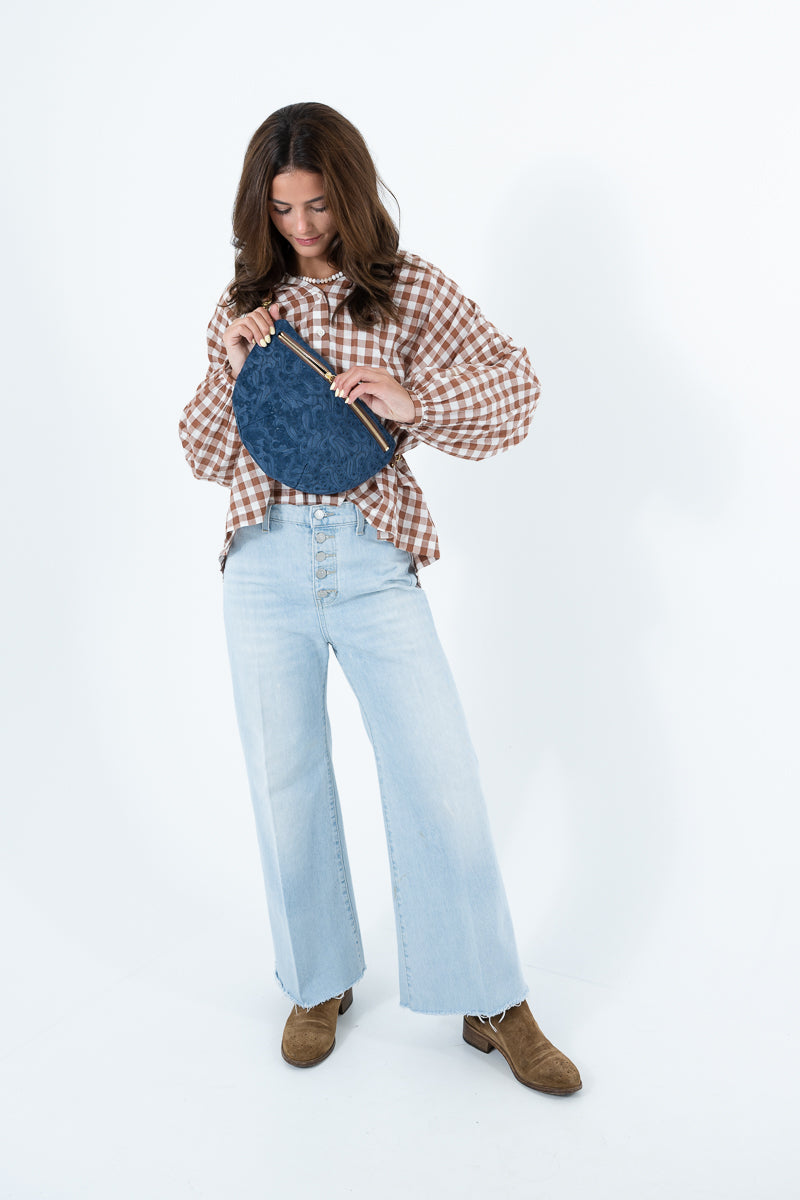 A woman modeling a brown and beige gingham shirt, light blue wide-leg jeans, and brown suede boots, holding a blue leather crossbody bag against a plain white studio background. Handcrafted by Meanwhile Back on the Farm in Lynchburg, Va.