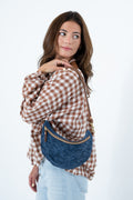 A woman wearing a brown and white checkered shirt and light blue jeans carries a blue crescent-shaped crossbody bag with gold hardware against a plain white background. Handcrafted by Meanwhile Back on the Farm in Lynchburg, Va.