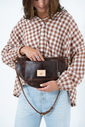 A woman wearing a brown and white gingham shirt and light blue jeans holds a dark brown leather belt bag with a small tan logo patch. The photo is cropped from the head down against a plain white background. Handcrafted by Meanwhile Back on the Farm in Lynchburg, Va.