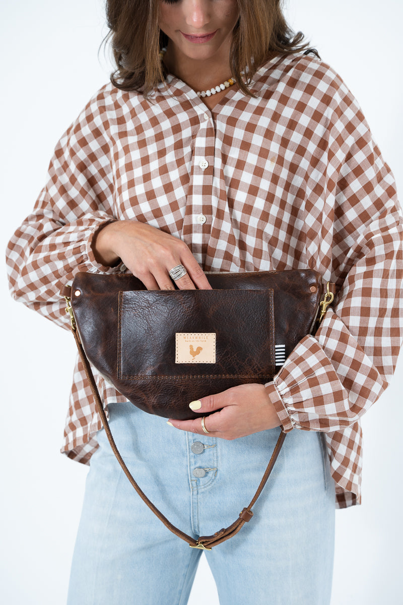 A woman wearing a brown and white gingham shirt and light blue jeans holds a dark brown leather belt bag with a small tan logo patch. The photo is cropped from the head down against a plain white background. Handcrafted by Meanwhile Back on the Farm in Lynchburg, Va.