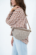 A woman wearing a brown and white checkered shirt and light blue jeans carries a taupe suede crescent-shaped crossbody bag with gold-tone hardware and a brown shoulder strap. Handcrafted by Meanwhile Back on the Farm in Lynchburg, Va.