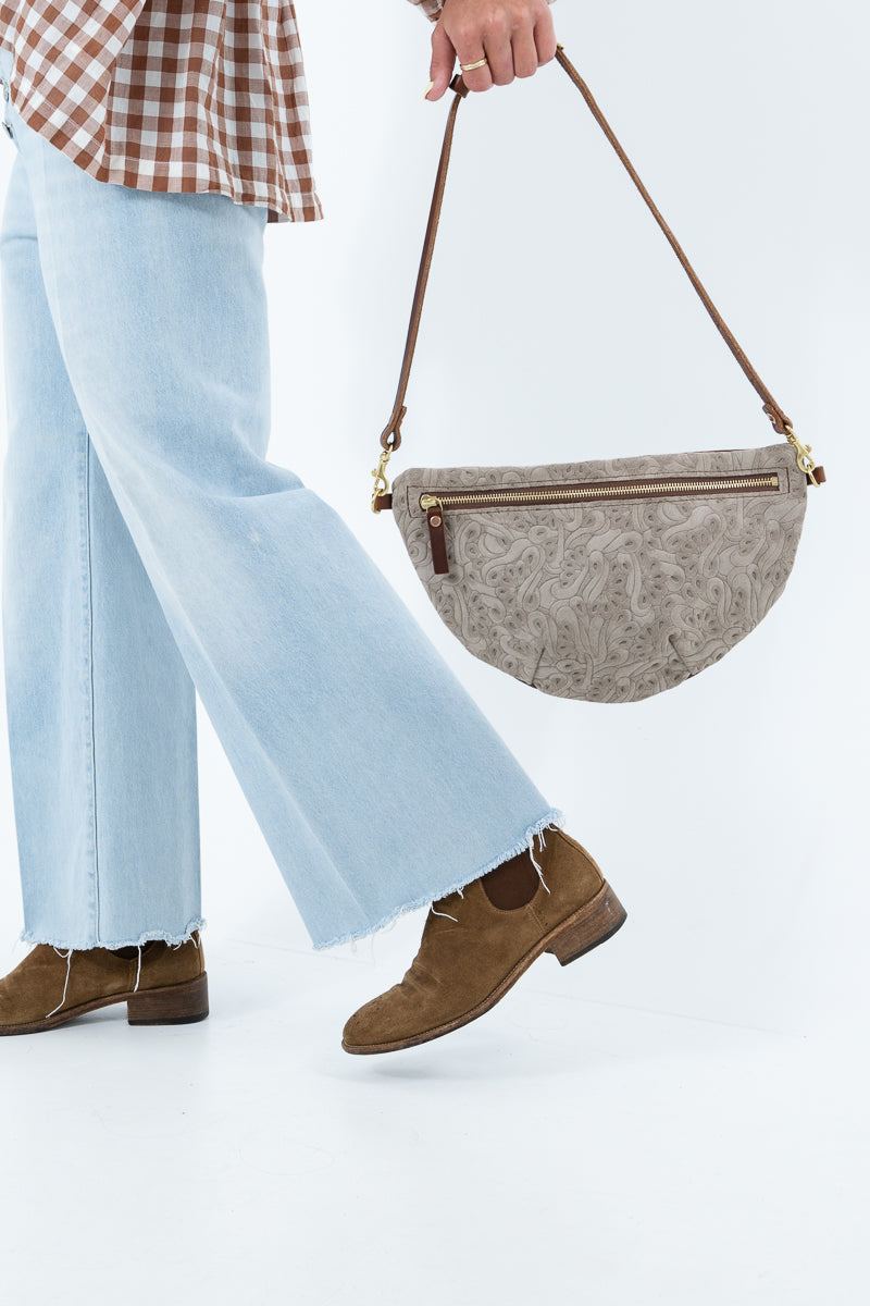 A person wearing light blue wide-leg jeans and brown suede shoes carries a taupe leather crescent-shaped shoulder bag with a brown strap and gold-tone hardware against a white studio background. Handcrafted by Meanwhile Back on the Farm in Lynchburg, Va.