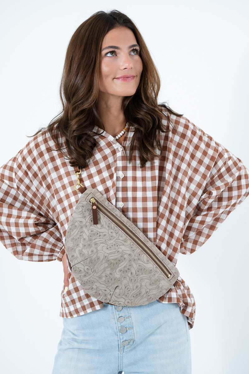 A woman wearing a brown and white gingham shirt and light blue jeans, carrying a taupe textured leather crescent crossbody bag with gold-tone hardware. Handcrafted by Meanwhile Back on the Farm in Lynchburg, Va.