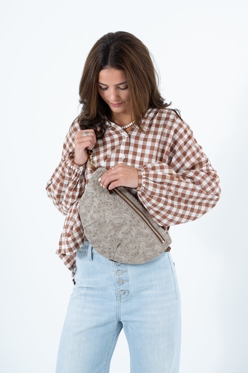 A woman wearing a brown and white gingham long-sleeve blouse and light blue jeans, holding a taupe textured crescent shoulder bag against a plain white background. Handcrafted by Meanwhile Back on the Farm in Lynchburg, Va.
